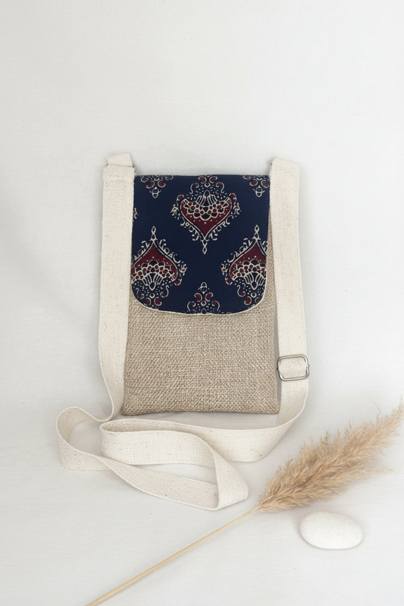 A small jute fabric crossbody bag with a witha  ajrakh patch work flap and a matching cream webbed cotton strap on a white background.